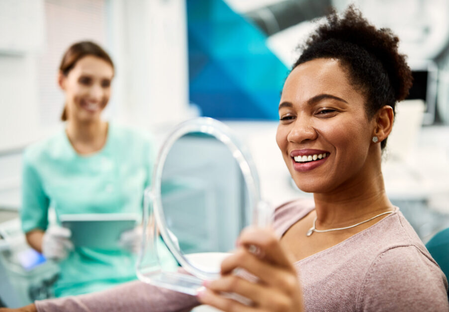 Happy black woman looking her teeth in a mirror after dental procedure at dentist's office.